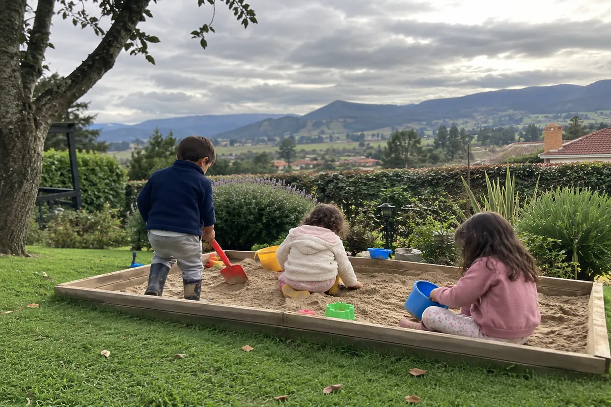 Dos o tres niños jugando en un arenero al aire libre con baldes y palas, vistos de lejos y de espaldas, bajo un cielo nublado de la Sabana