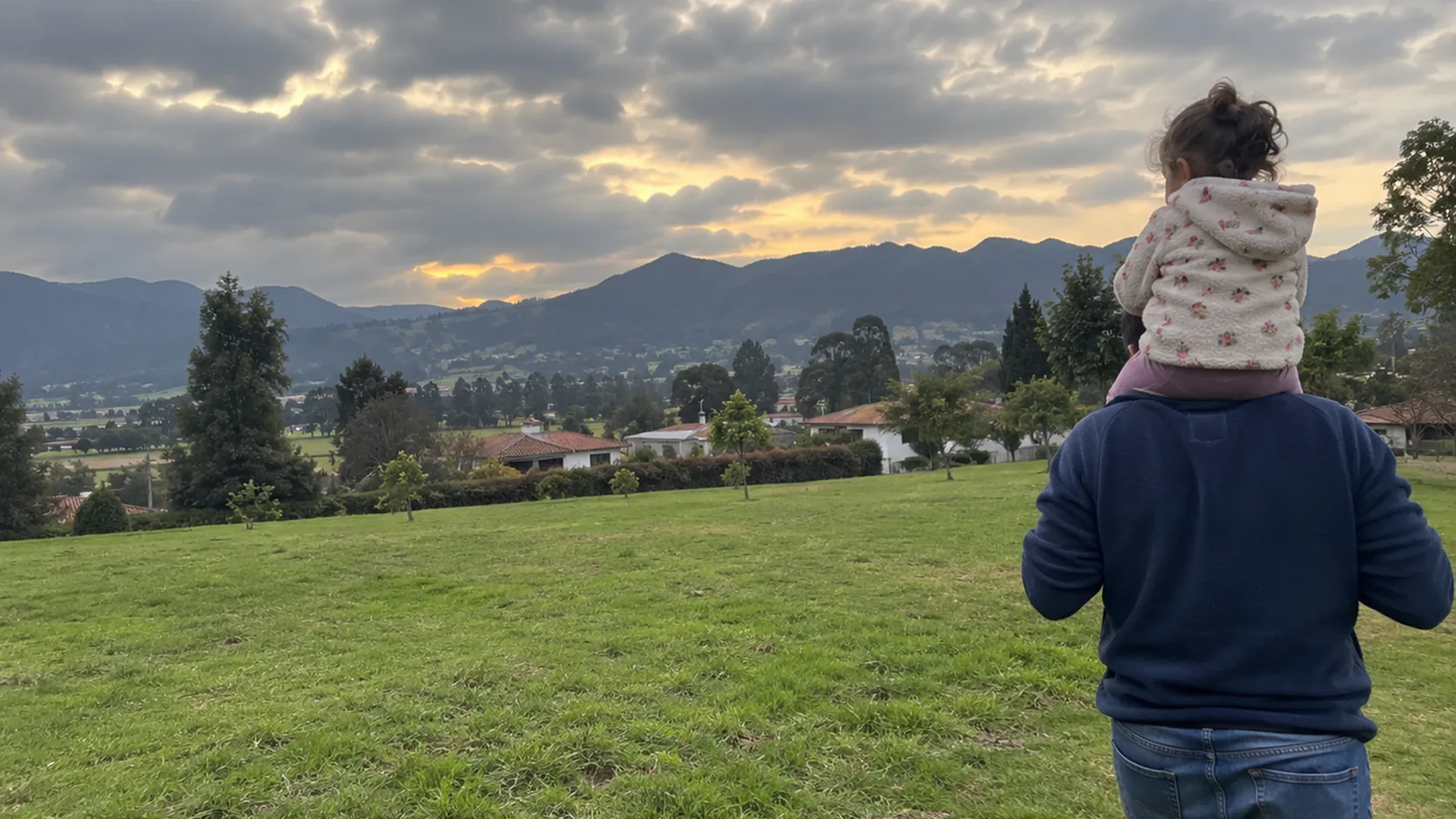 Un papá lleva en hombros a su hijo pequeño por un parque bajo el cielo nublado de la Sabana