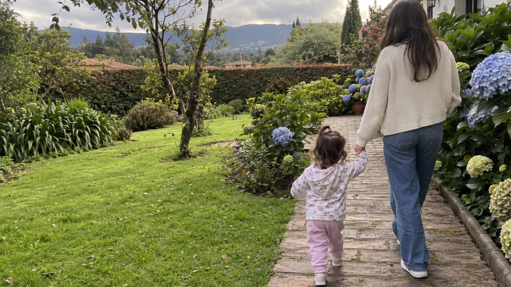 Una niña de dos años camina de la mano de su mamá por un sendero con hortensias