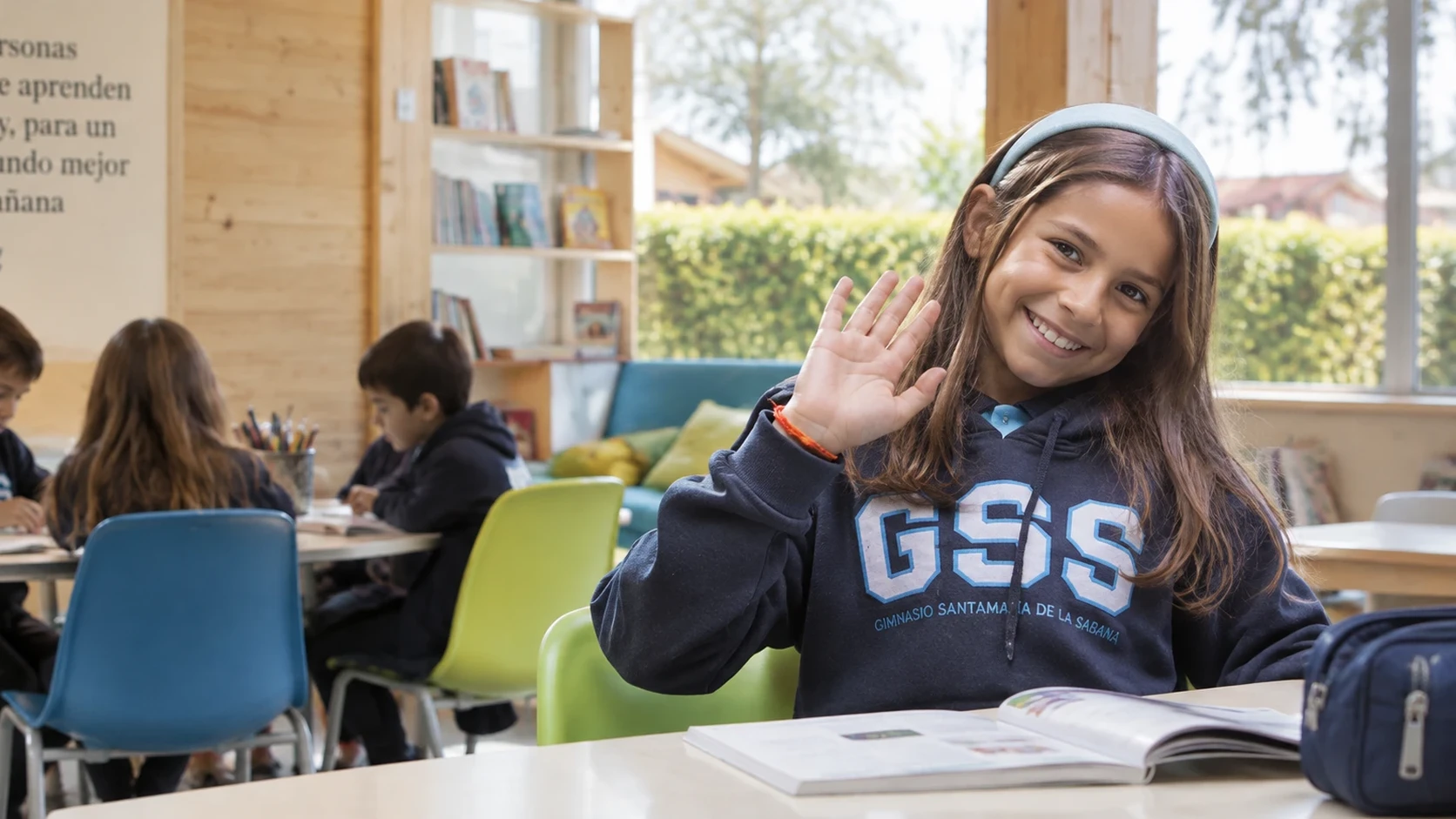Estudiante de primaria saludando con la mano desde su mesa en la biblioteca del colegio